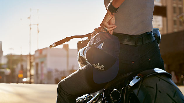 In the close-up, a motorcyclist has the Boxer cap from the collection in her hand and is wearing the RoadCrafted jeans for women.