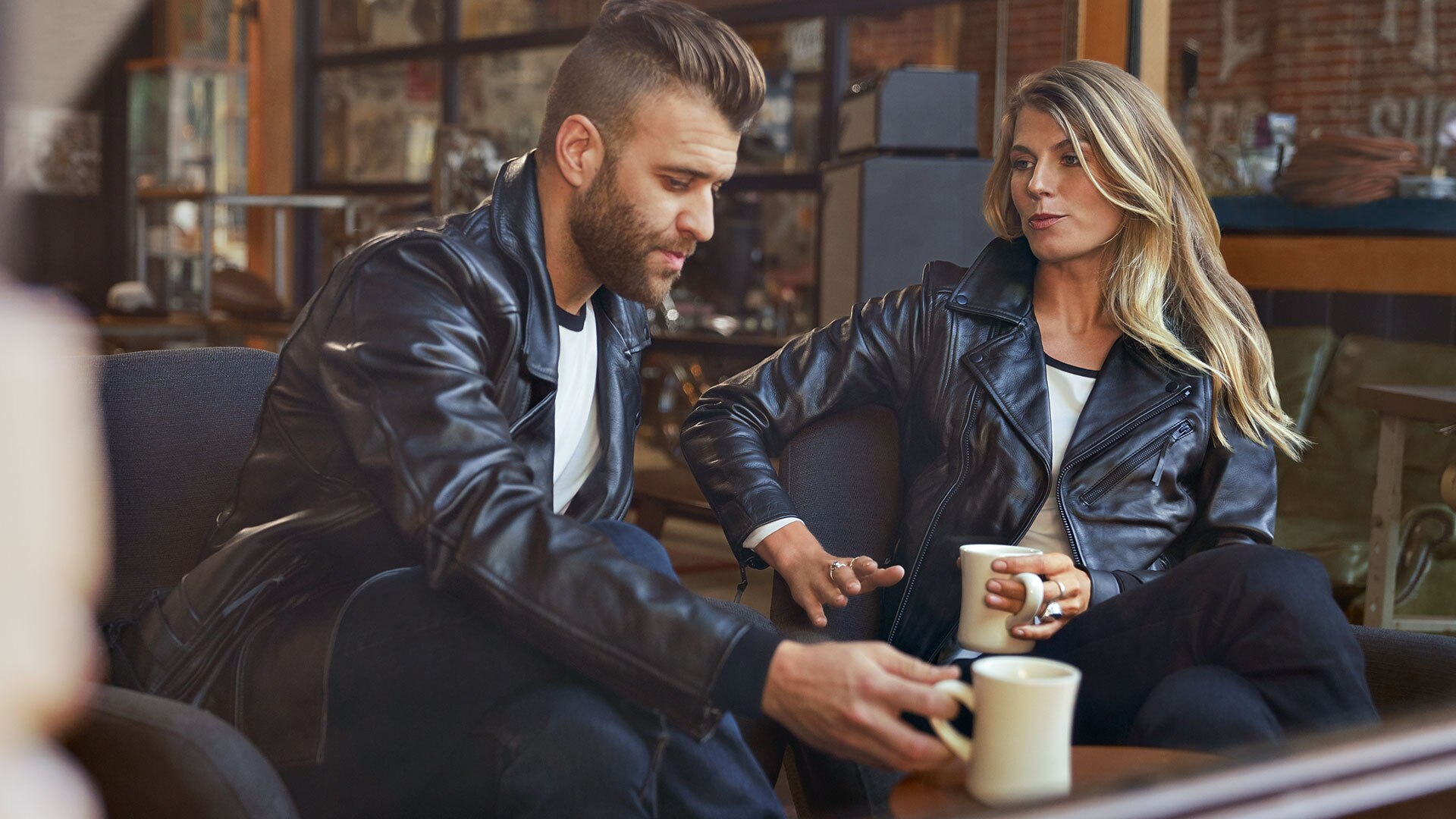 In the image sit a young man in the men’s FlatTwin leather jacket and a young woman in the women's FlatTwin leather jacket across from him.