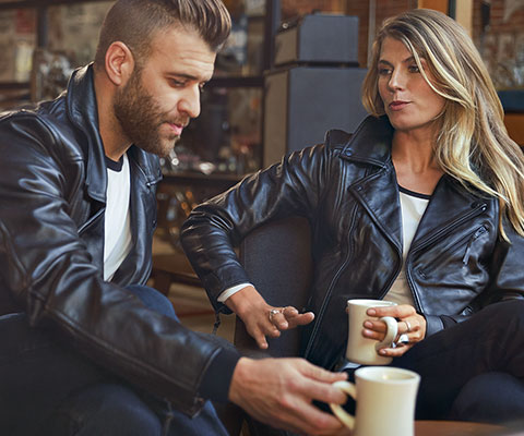 In the image sit a young man in the men’s FlatTwin leather jacket and a young woman in the women's FlatTwin leather jacket across from him.