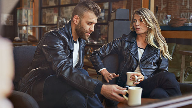 In the image sit a young man in the men’s FlatTwin leather jacket and a young woman in the women's FlatTwin leather jacket across from him.